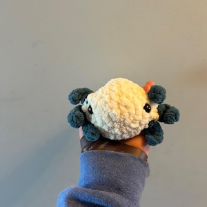 Może przedstawiać: Pluszowa zabawka w kształcie aksolotla, wykonana na szydełku. Zabawka jest biała z niebieskimi, pierzastymi skrzelami i czarnymi oczami.