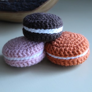 May include: Three crocheted macarons in brown, pink, and orange. Each has a white filling. The macarons are stacked together. The background is a light, neutral colour. The macarons are handmade.