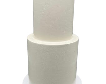 May include: A two-tiered white cake with smooth frosting. The cake is on a white cardboard cake base.