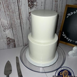 May include: A two-tiered white frosted cake on a clear cake stand. A cake server and knife are on the table. The cake is next to a chalkboard sign with a floral design. The image includes the text "Create Something From Jenn With Love."