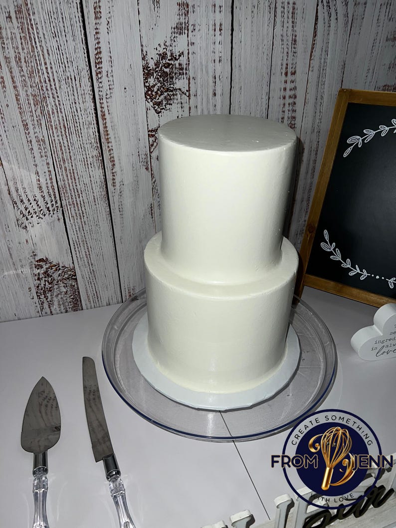 May include: A two-tiered white frosted cake on a clear cake stand. A cake server and knife are on the table. The cake is next to a chalkboard sign with a floral design. The image includes the text "Create Something From Jenn With Love."