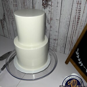 May include: A two-tiered white frosted cake on a clear plastic cake stand. The cake is round and has a smooth, even surface. A cake knife is visible on the left. The cake is on a white surface with a wooden background.