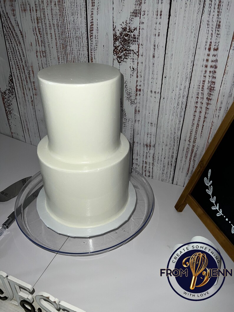 May include: A two-tiered white frosted cake on a clear plastic cake stand. The cake is round and has a smooth, even surface. A cake knife is visible on the left. The cake is on a white surface with a wooden background.