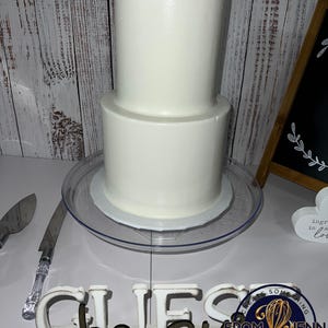 May include: A two-tiered white cake on a clear cake stand. A cake knife with a clear handle and a silver blade is to the left. A wooden sign spells out "CHEFS" in white letters with brown accents. A small sign reads "ingredients is love".