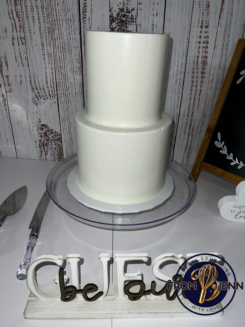 May include: A two-tiered white cake on a clear cake stand. A cake knife with a clear handle and a silver blade is to the left. A wooden sign spells out "CHEFS" in white letters with brown accents. A small sign reads "ingredients is love".