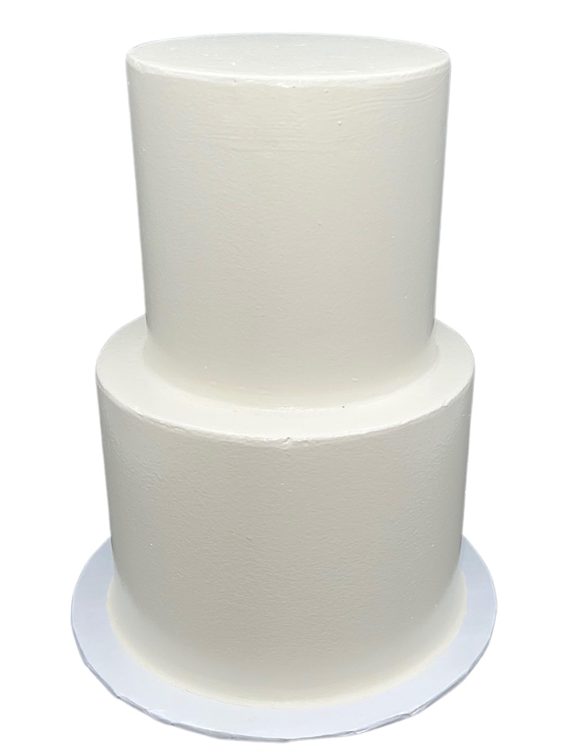 May include: A two-tiered white cake with a smooth, frosted finish. The cake sits on a white, round cake board. The top tier is smaller than the bottom tier, creating a classic layered look. The cake is a neutral color, suitable for various celebrations.