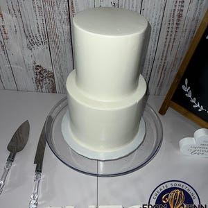 May include: A two-tiered white frosted cake on a clear glass cake stand. Two cake serving utensils with clear handles are to the left. A wooden sign with the word "Bless" is in the foreground.
