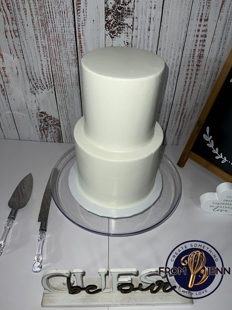 May include: A two-tiered white frosted cake on a clear glass cake stand. Two cake serving utensils with clear handles are to the left. A wooden sign with the word "Bless" is in the foreground.
