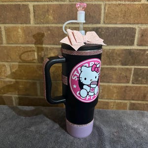 May include: Black travel mug with a pink Hello Kitty design and a pink glitter lid. The mug has a handle and a pink glitter band around the bottom.