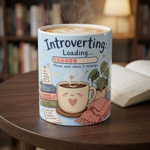 May include: A light blue ceramic mug featuring the text Introverting: Loading... Please wait while I recharge. The design includes a stack of books, a potted plant, a pink knitted blanket, and a smiling coffee cup with a heart graphic.