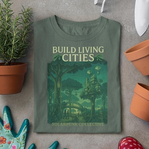 Könnte beinhalten: Grünes T-Shirt mit einem Grafikdruck einer Baumhausstadt und dem Text "Build Living Cities" und "Solarpunk Collective".