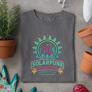 May include: A dark gray t-shirt with a green, pink, and blue graphic of a city skyline inside a sun with the text "Solarpunk Collective - Powered by Sun - Fueled by Hope".