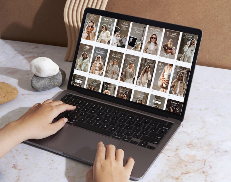 May include: A laptop displaying a grid of social media posts with images of a woman in various outfits. The laptop is open, with hands typing on the keyboard. The background includes decorative stones and a wooden element.
