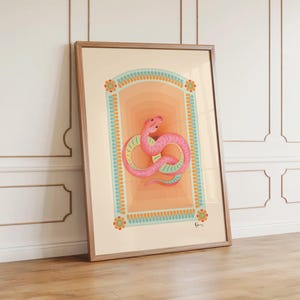 Puede incluir: Una impresión artística enmarcada que presenta una serpiente rosa estilizada en un arco decorativo. La serpiente tiene detalles en amarillo y verde. El arco tiene un fondo degradado naranja claro, enmarcado por un borde turquesa con detalles florales. El marco es de color marrón claro.