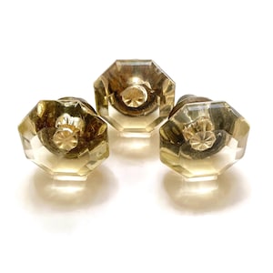 May include: Three vintage octagonal glass cabinet knobs with brass hardware. The knobs have a clear, faceted design with a yellowish tint, and are arranged against a white background. These decorative knobs are suitable for furniture restoration or DIY projects.