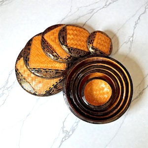 May include: A set of round, nested containers featuring a woven light brown bamboo center and a dark brown lacquered border decorated with gold floral patterns. The set includes several circular lids with matching woven bamboo and floral designs.