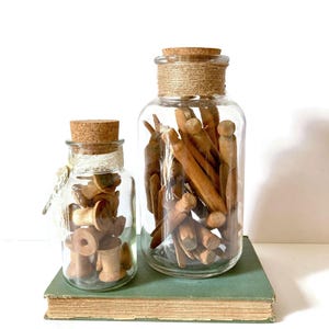 May include: Two clear glass jars with cork stoppers, one small and one large, filled with wooden spools and clothespins. The larger jar has a twine-wrapped neck. The jars sit on a vintage green book, creating a rustic, vintage aesthetic.