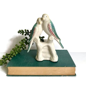 May include: A vintage ceramic figurine of two birds perched on a white tree branch. The birds have white bodies with teal and red accents on their wings. The figurine is displayed on a green book.