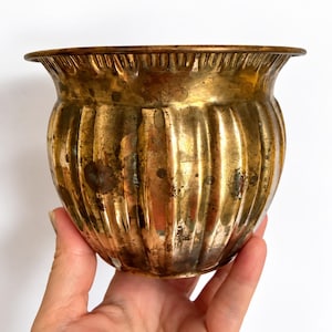 May include: A round, metallic brass planter featuring a vertical ribbed texture along its body and a wide, flared rim. The surface displays a weathered, aged patina with dark spots and variations in the golden-brown metal finish.