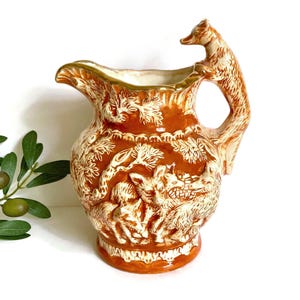 May include: A brown and cream-colored ceramic pitcher with a fox-shaped handle. The pitcher features a relief design of foxes and foliage. The rim and base have a cream-colored trim. The pitcher is a decorative item.