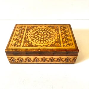 May include: A rectangular wooden box with intricate floral and scrollwork carvings. The box features a central floral design on the lid, surrounded by a border of detailed patterns. The wood has a warm, natural tone with darker edges.
