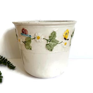 May include: Off-white ceramic planter with a whimsical design. The planter features a raised relief pattern of daisies, green leaves, and small creatures like a snail and a butterfly. The rim is slightly flared, and the overall shape is cylindrical.