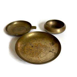 May include: Three brass vessels consisting of a small round bowl, a shallow dish with a side handle, and a larger circular tray featuring an etched floral pattern in the center. Each piece has a metallic gold finish with visible signs of aging.