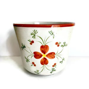 May include: A white ceramic planter with a hand-painted floral design. The planter features a red rim and a central flower motif in shades of orange and yellow, surrounded by green leaves and red berries. The planter is suitable for indoor or outdoor use.