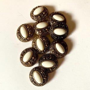 May include: A collection of antique brass-toned buttons with oval, off-white centers. The buttons have an ornate, filigree design around the edges. The buttons are arranged on a white surface.