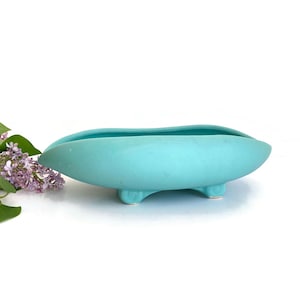 May include: A turquoise ceramic planter with four feet. The planter is shaped like a boat and has a smooth, glossy finish.