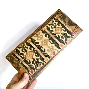 May include: Rectangular box with a patterned design in black, tan, and red. The box has a gold trim and a decorative pattern on the top. The box is held by a hand.