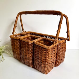 May include: A brown woven wicker caddy featuring four rectangular compartments and a tall, arched handle. The basket is constructed with a tight, uniform weave pattern, providing a structured and open design for storage and organization.
