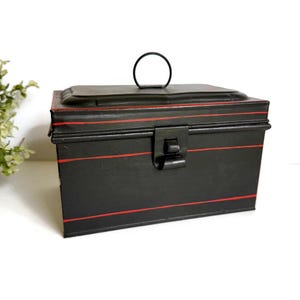 May include: A rectangular black metal storage box featuring thin horizontal red stripes along the lid and front panel. The box includes a hinged lid with a circular metal handle on top and a central metal latch mechanism on the front face.