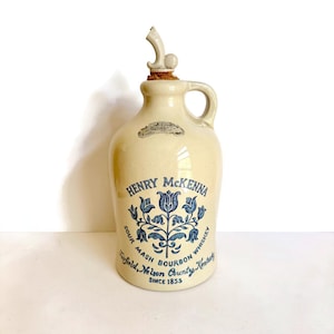 May include: Cream-colored ceramic jug with a handle and a decorative stopper. The jug features blue floral designs and text that reads "Henry McKenna Sour Mash Bourbon Whiskey, Fairfield, Nelson County, Kentucky, Since 1855."