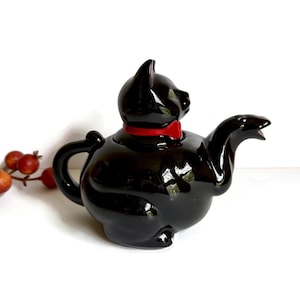 May include: A glossy black ceramic teapot shaped like a seated cat. The teapot features a bright red collar around the neck, a curved handle on the left side, and a spout on the right side that resembles an open mouth. The surface is reflective.
