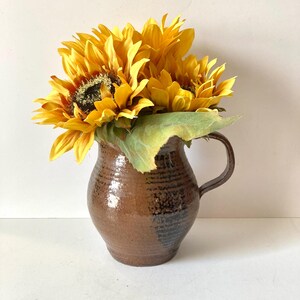 May include: A brown ceramic pitcher with a handle, filled with bright yellow sunflowers. The sunflowers have dark centers and green leaves. The pitcher has a textured surface with horizontal lines.