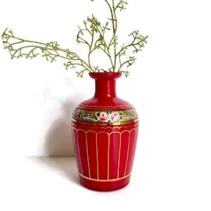 May include: A vibrant red vase with gold accents and a floral design. The vase has a narrow neck and a wider base, with vertical lines adding texture. Green sprigs are placed inside the vase.