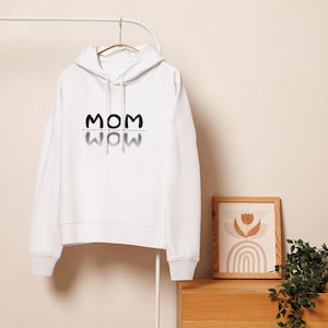 Puede incluir: Sudadera con capucha blanca con la palabra "MOM" en negro, con un efecto de sombra reflejada debajo. La sudadera con capucha está colgada de una percha de metal. Una imagen enmarcada en madera y una pequeña planta están en el fondo.
