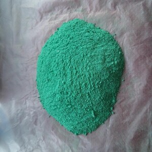 May include: A pile of bright green powder on a white surface. The powder is finely ground and has a uniform texture.