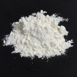 May include: A close-up image of a heap of white powder on a black surface. The powder is finely ground and has a slightly uneven texture. The lighting accentuates the irregular form of the powder.