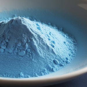 May include: A close-up of a pile of light blue powder in a white bowl. The powder is fine and smooth, and it has a slightly grainy texture.