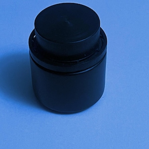 Può includere: Un barattolo di plastica nero con un coperchio nero. Il barattolo è chiuso.