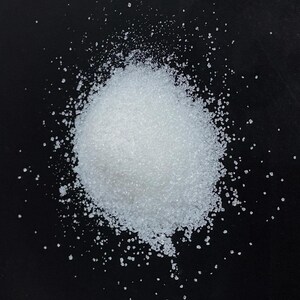 Pode incluir: Uma imagem em close-up de um monte de grânulos cristalinos brancos sobre um fundo preto. Os grânulos estão espalhados ao redor do monte principal, criando uma aparência texturizada. A imagem destaca a textura granular dos cristais.