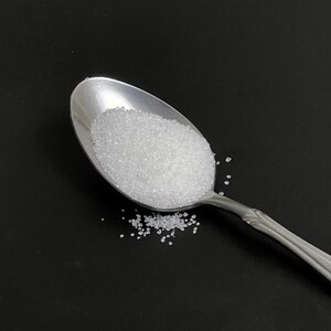 以下が含まれることがあります： 黒い背景に白い砂糖がたっぷり入った銀のスプーン