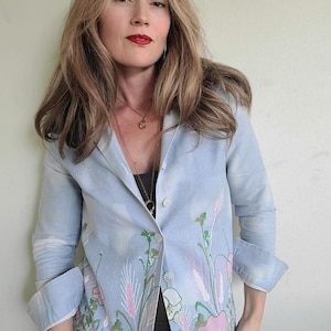 May include: A light blue blazer with a floral design featuring pink and white flowers, green stems, and light purple accents. The blazer has a button closure and rolled-up sleeves.