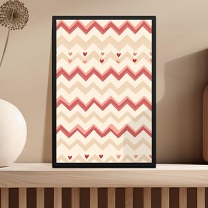 Puede incluir: Una impresión artística enmarcada con un borde negro que presenta un patrón de chevrón repetido en tonos rojos y beige. Pequeños corazones rojos están dispersos por el diseño. La obra de arte se exhibe en una estantería de madera.