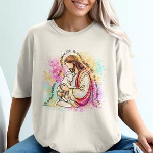 Puede incluir: Camiseta color crema con una ilustración lineal de Jesús sosteniendo un cordero, con el texto "He is risen Matthew 28:6" en un patrón circular. El diseño incluye salpicaduras de color estilo acuarela.
