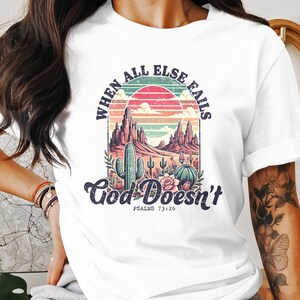 May include: White t-shirt featuring a desert landscape design with cacti, mountains, and a sunset. The text "When All Else Fails God Doesn't" is arched above the image, with "Psalms 73:26" below. A casual, comfortable tee.