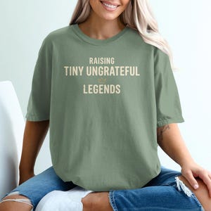 以下が含まれることがあります： ライトベージュの「RAISING TINY UNGRATEFUL LEGENDS」の文字が入ったセージグリーンのTシャツ。 ゆったりとしたフィット感のシャツが着用されています。