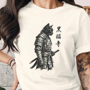 Peut inclure: T-shirt crème avec une illustration noire d'un chat en armure de samouraï, tenant un katana. Des caractères japonais sont imprimés à droite. Le chat est détaillé avec une armure complexe et une expression sévère.